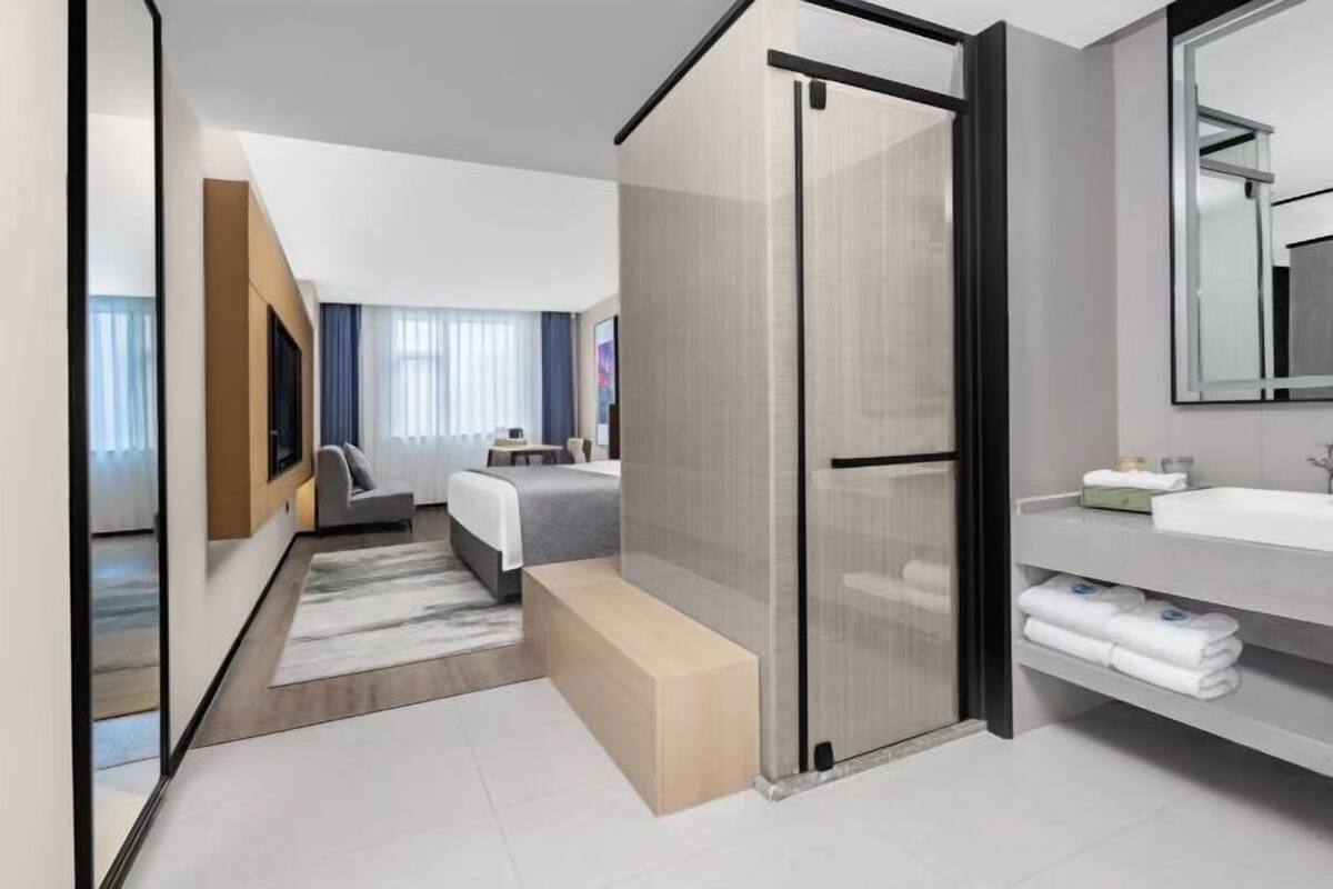 Quarto de hotel moderno com cama, um banco, banheiro com pia e porta-toalhas, e um espelho grande.