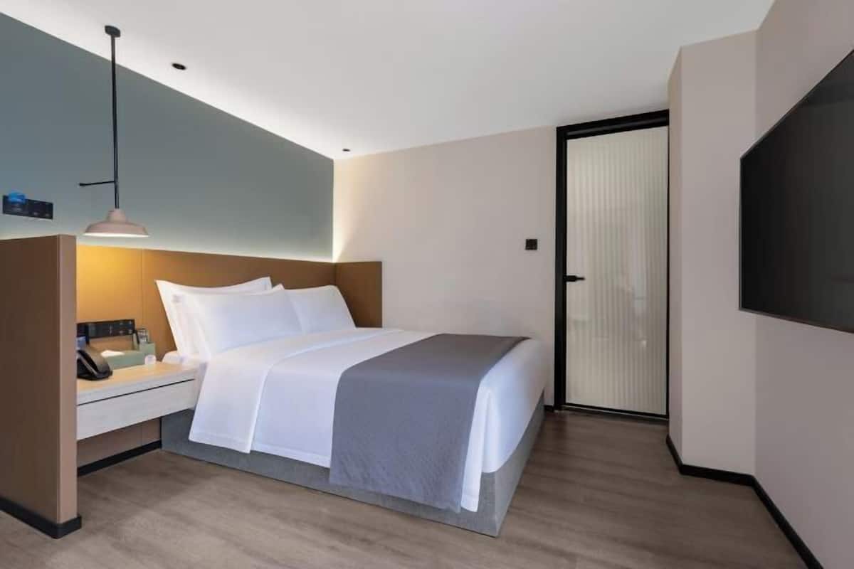 Quarto de hotel moderno com cama, mesa de cabeceira, abajur e televisão fixada na parede.