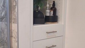 Un baño con pared de mármol, un mueble blanco con cajones y estantes, una planta y artículos de aseo.