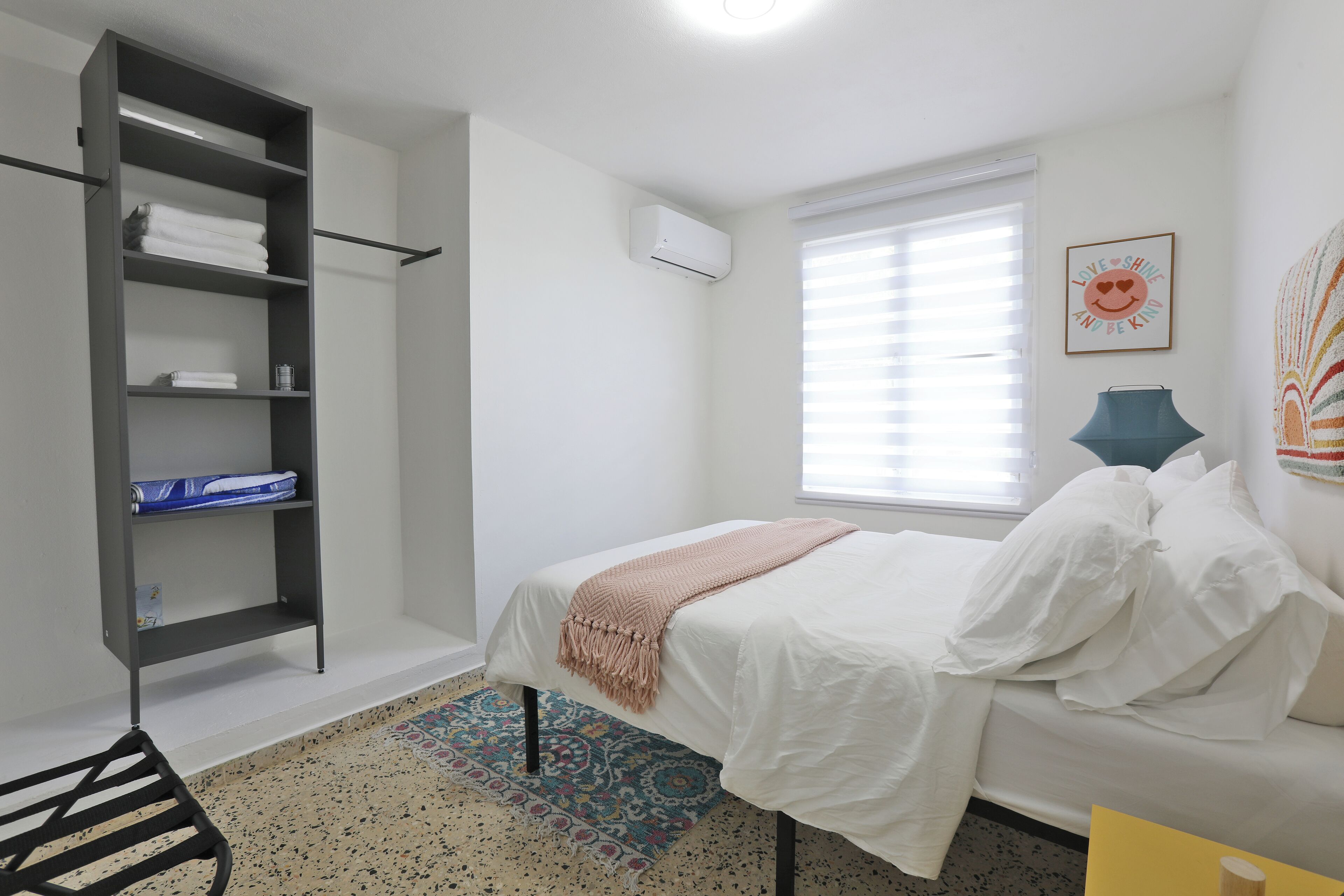 A bedroom with a bed, a wardrobe, a window with blinds, a wall-mounted air conditioner, and a decorative wall piece.