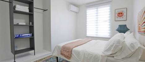 A bedroom with a bed, a wardrobe, a window with blinds, a wall-mounted air conditioner, and a decorative wall piece.