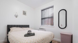 A neatly made bed with a patterned bedspread, a bedside table, a wall-mounted mirror, and a window with blinds. - Roof Terrace | W/M | Smart TV | Free Parking (Old Swan)