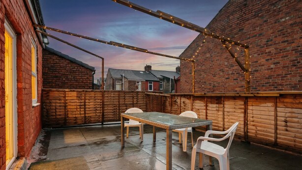 An outdoor patio area with a glass-top table and white plastic chairs, surrounded by wooden walls and a brick building in the background. - Roof Terrace | W/M | Smart TV | Free Parking (Old Swan)