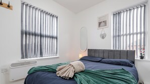 A bedroom with a bed, a radiator, a window with blinds, a wall-mounted mirror, and a framed picture.