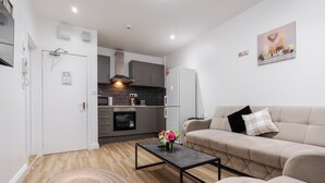 A modern living room with a beige sofa, a coffee table with a vase of flowers, and a kitchen in the background.