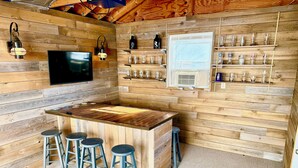 A wooden interior with a bar area, stools, a TV, and shelves with glasses.
