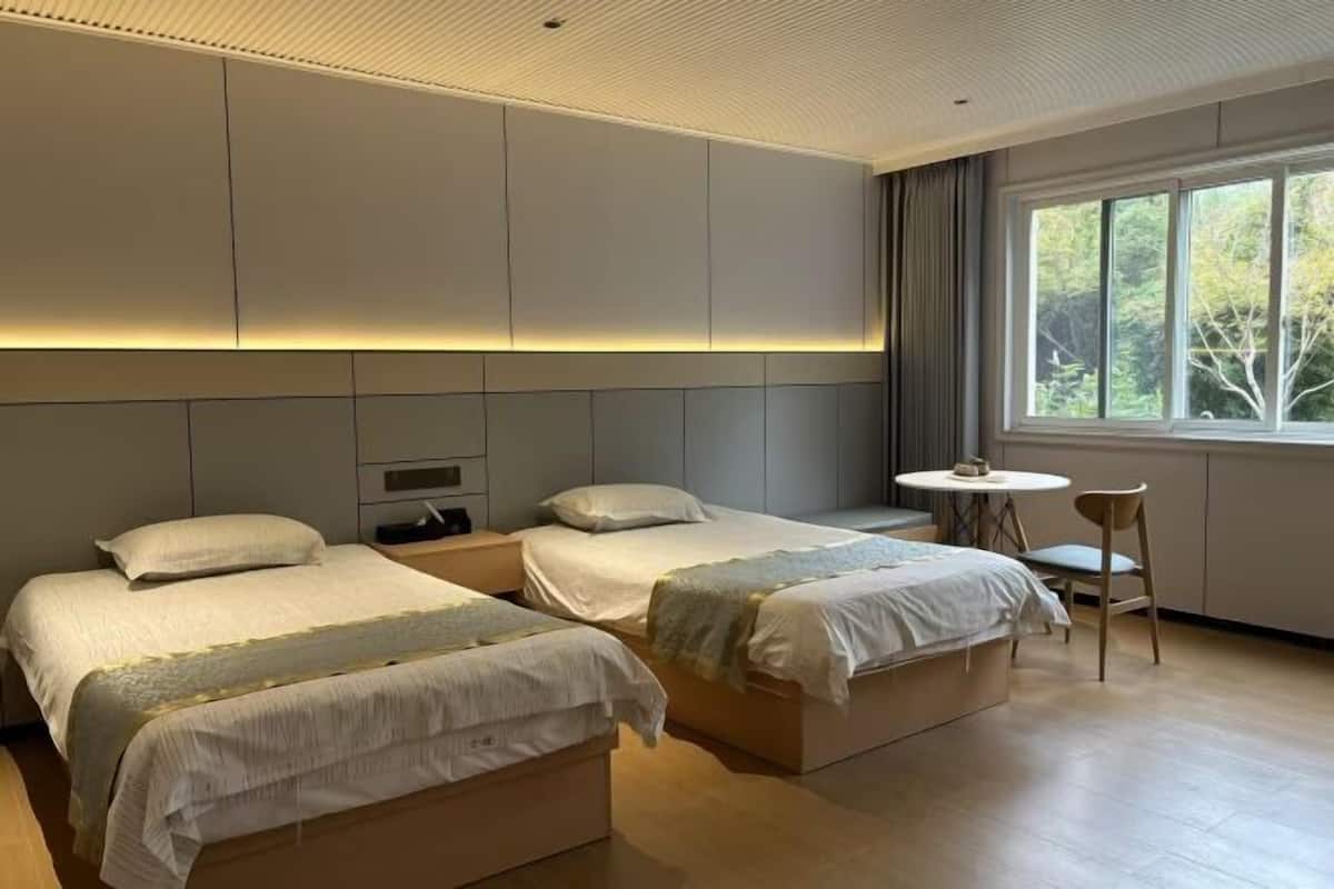 A hotel room with two beds, a small table, a chair, and a window with a view of trees.