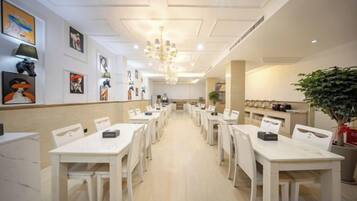 A hotel dining area with white tables and chairs, a chandelier, and artwork on the walls.