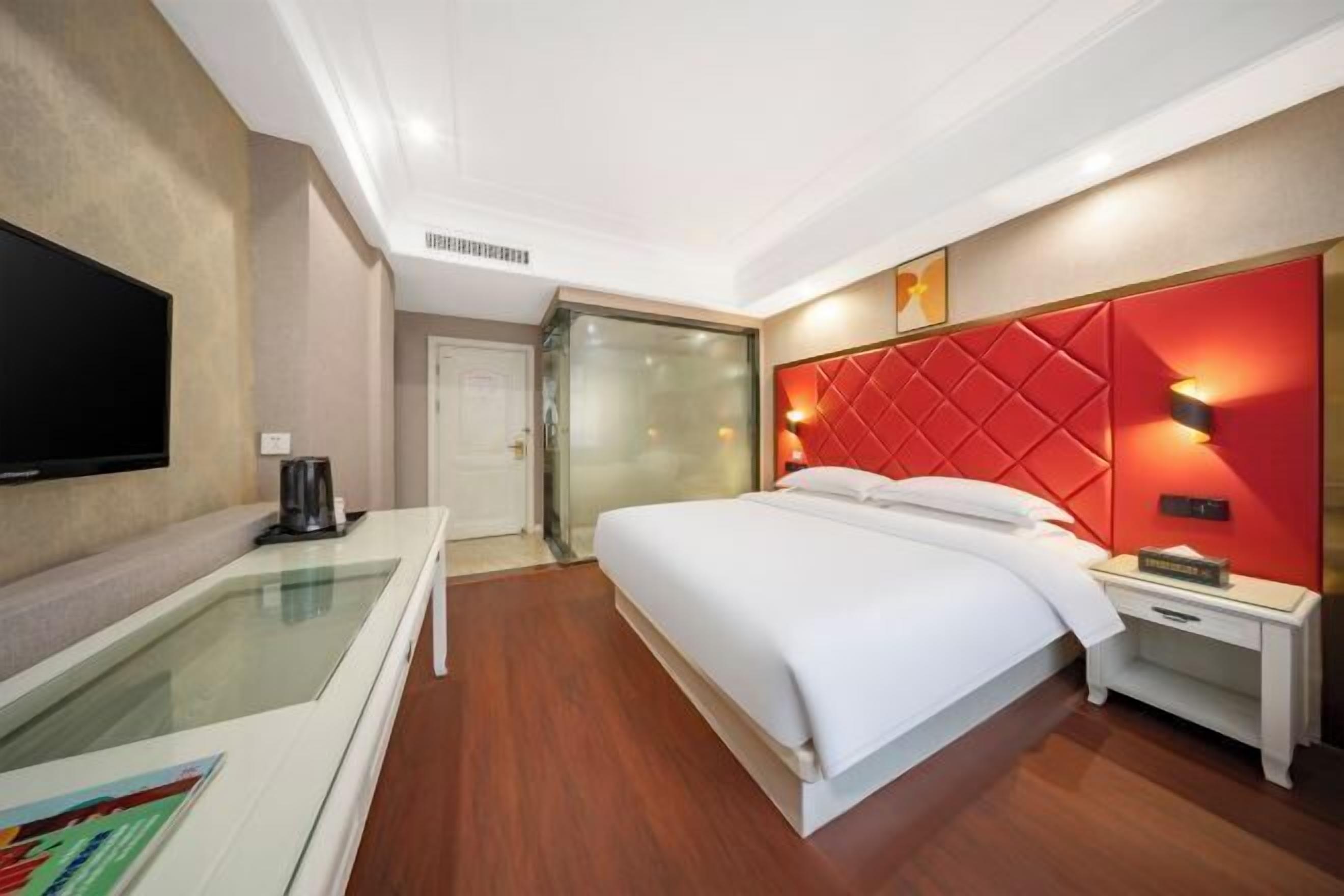 Habitación de hotel con cama, mesita de noche, televisión y separador de vidrio.