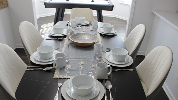 Een eettafel gedekt voor zes personen, met witte borden, kommen en bekers, omringd door witte fauteuils.
