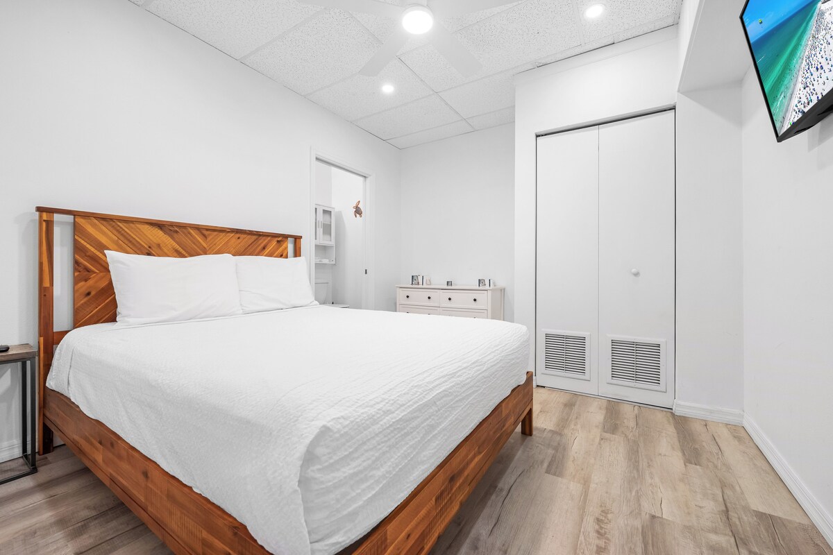 Une chambre à coucher avec un lit en bois, du linge de lit blanc, une armoire blanche et un téléviseur fixé au mur.