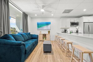 A modern living room with a blue sofa, a grey ottoman, wooden stools, a kitchen with white cabinets, a microwave, and a refrigerator. - Mallory Squared (Siesta Key)