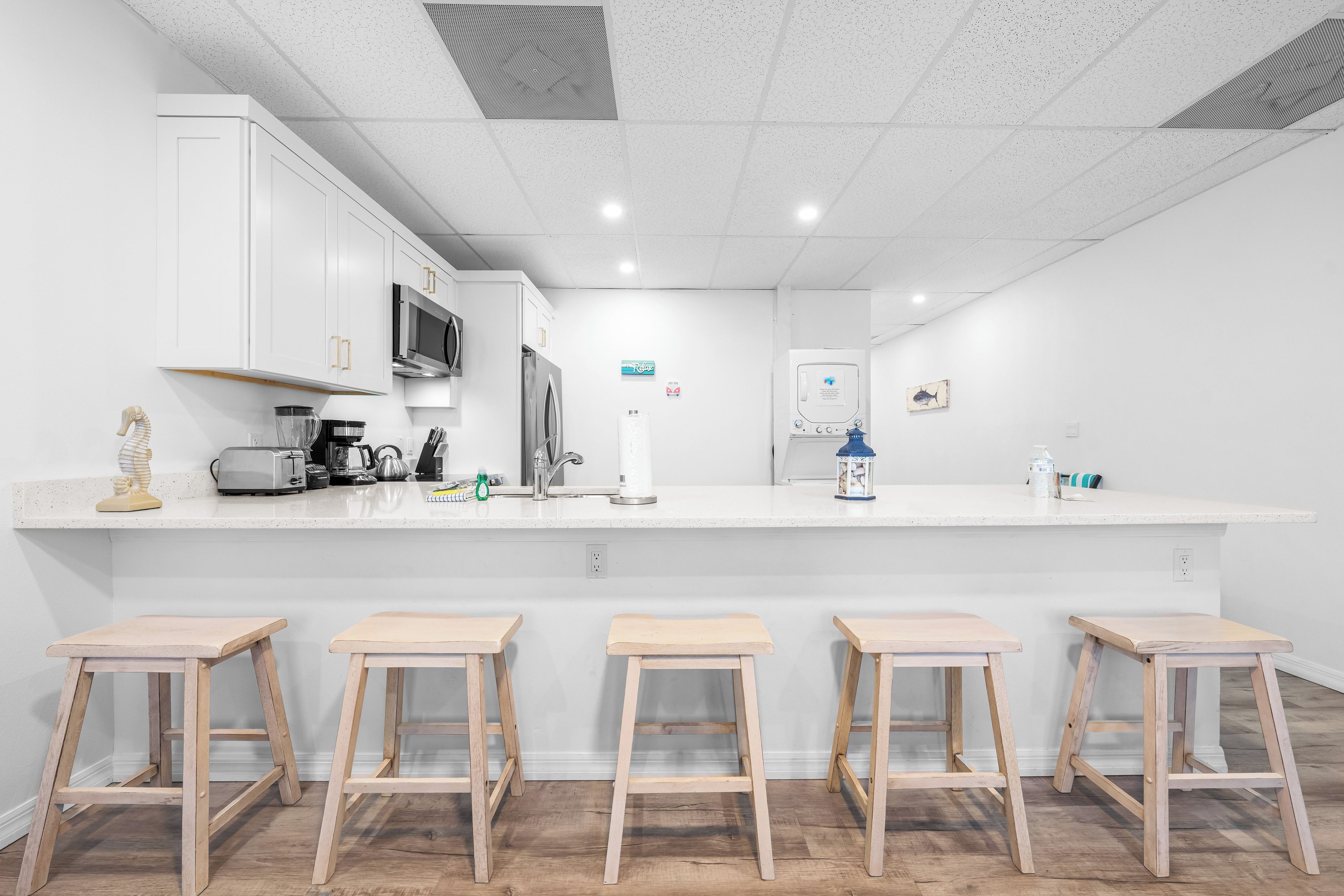 A modern kitchen with white cabinetry, a microwave, a refrigerator, and a sink. There are four wooden stools at the counter.