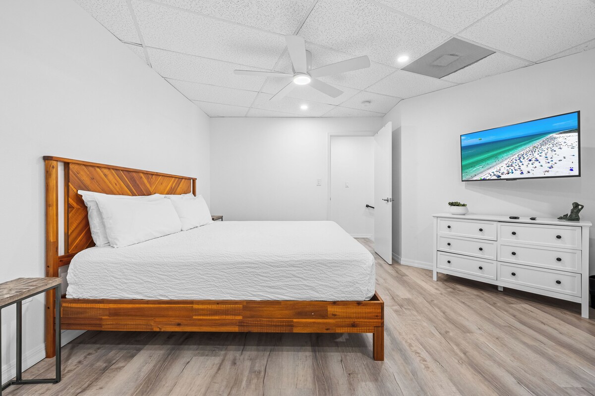 Une chambre avec un lit en bois, du linge de lit blanc, un ventilateur de plafond, un téléviseur fixé au mur et une commode blanche.