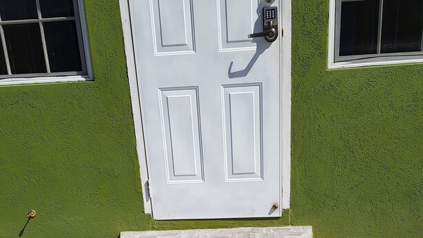 A set of stone steps leading to a white door with a keypad lock, set against a green wall with two windows.