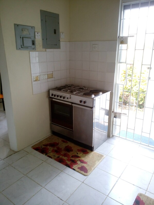 A kitchen with a gas stove, oven, and tiled walls. There is a door with a metal gate and a window with a view of greenery outside.
