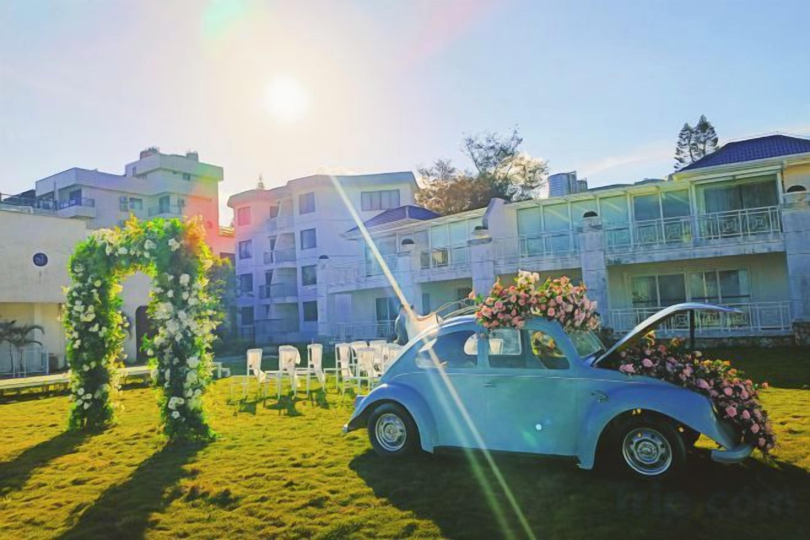 A vintage car with a floral arrangement, a grassy area, and a building with balconies in the background.
