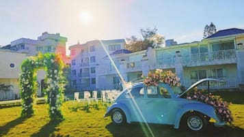 A vintage car with a floral arrangement, a grassy area, and a building with balconies in the background.