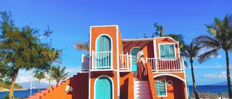 A two-story building with a staircase leading to a balcony, surrounded by palm trees and a clear blue sky.