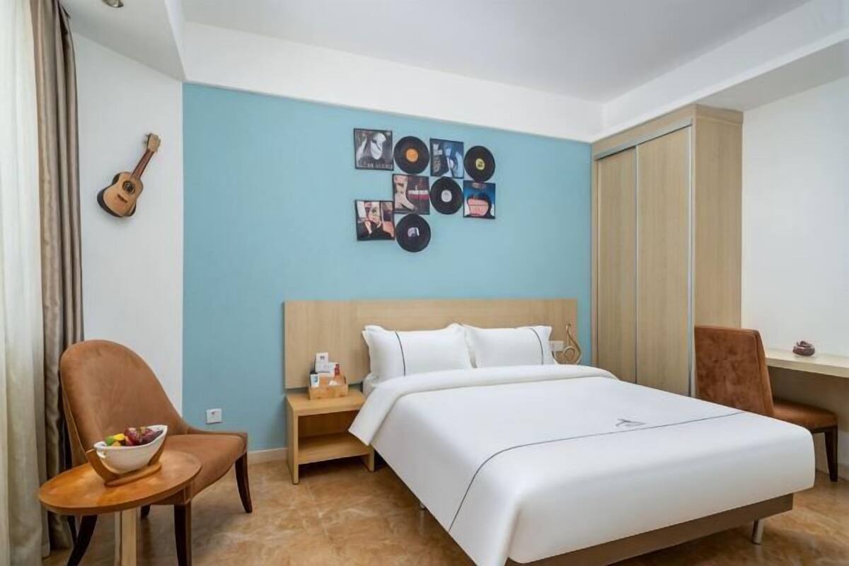 Un dormitorio con una cama, una silla, una mesita de noche, una guitarra y una pared decorada con portadas de discos.