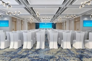 Uma sala de conferências com fileiras de cadeiras brancas, palco com fundo azul e piso coberto de carpete.