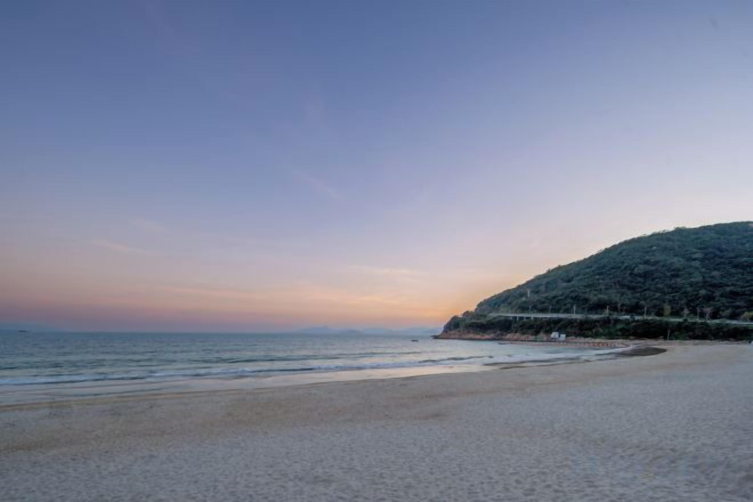 A beach with a gentle slope leading to the water, a clear sky, and a distant hill with vegetation.