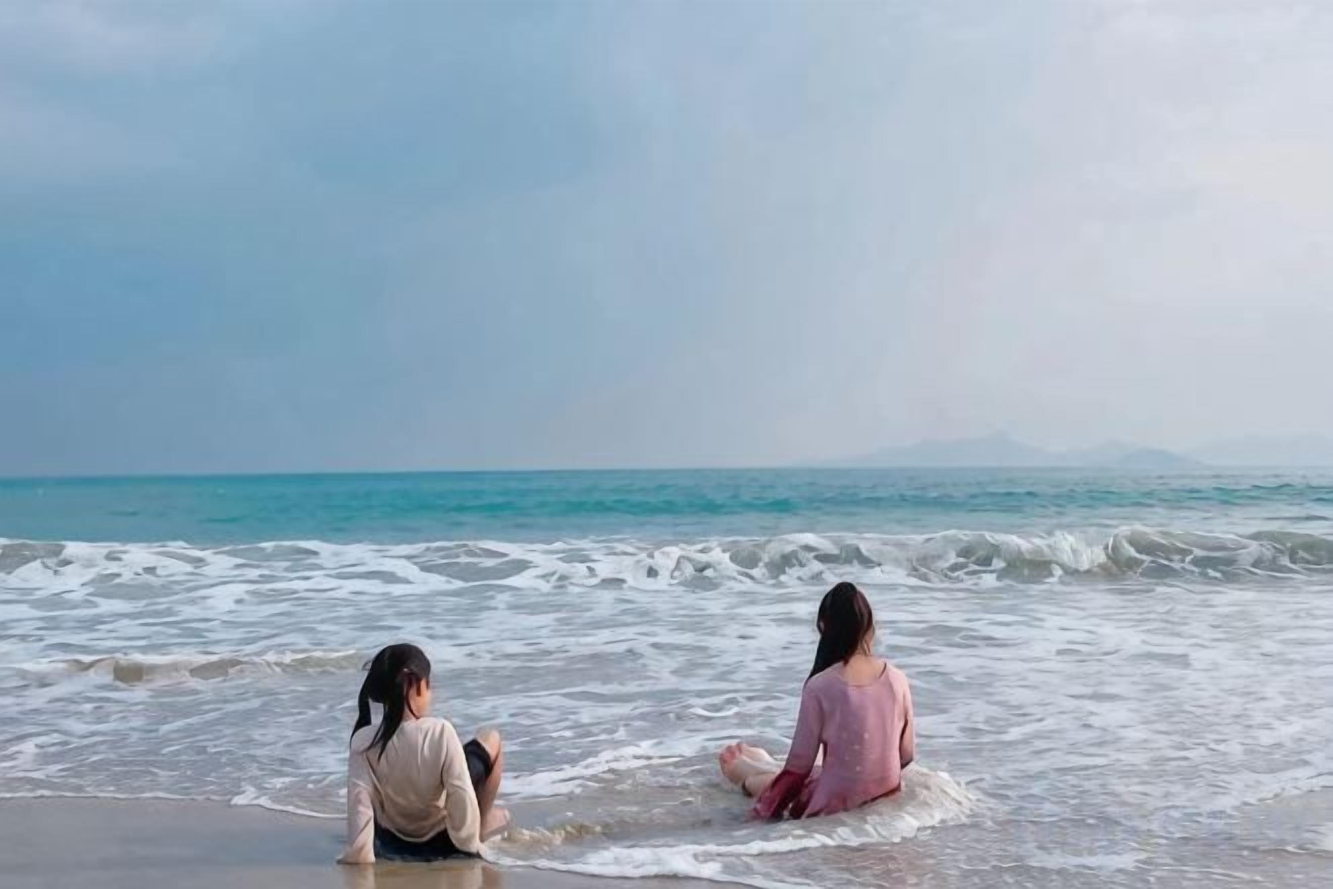 Two individuals sitting on a beach with the ocean in the background.