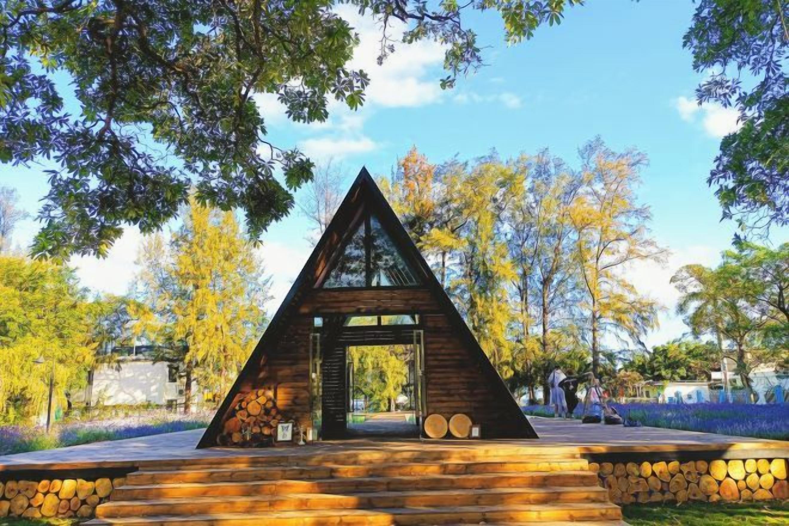Une structure triangulaire en bois, dotée d’une porte, entourée d’arbres et s’ouvrant sur un ciel dégagé.