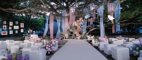 An outdoor wedding venue with a white aisle, floral arrangements, and a backdrop of trees.