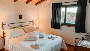 A bedroom with a wooden ceiling, a bed with white bedding, a nightstand with a lamp, a window with curtains, and a chair with a table. - Mondifeso Chalet (Etna+vineyard+jacuzzi) (Pedara)