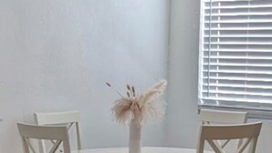 Un comedor con una mesa redonda y cuatro sillas, un jarrón con flores secas, y una ventana con persianas.