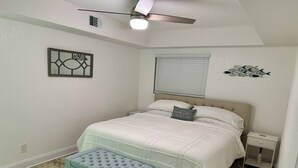 Un dormitorio con cama, mesita de noche, banqueta, ventilador de techo, ventana con persianas y decoración en la pared.