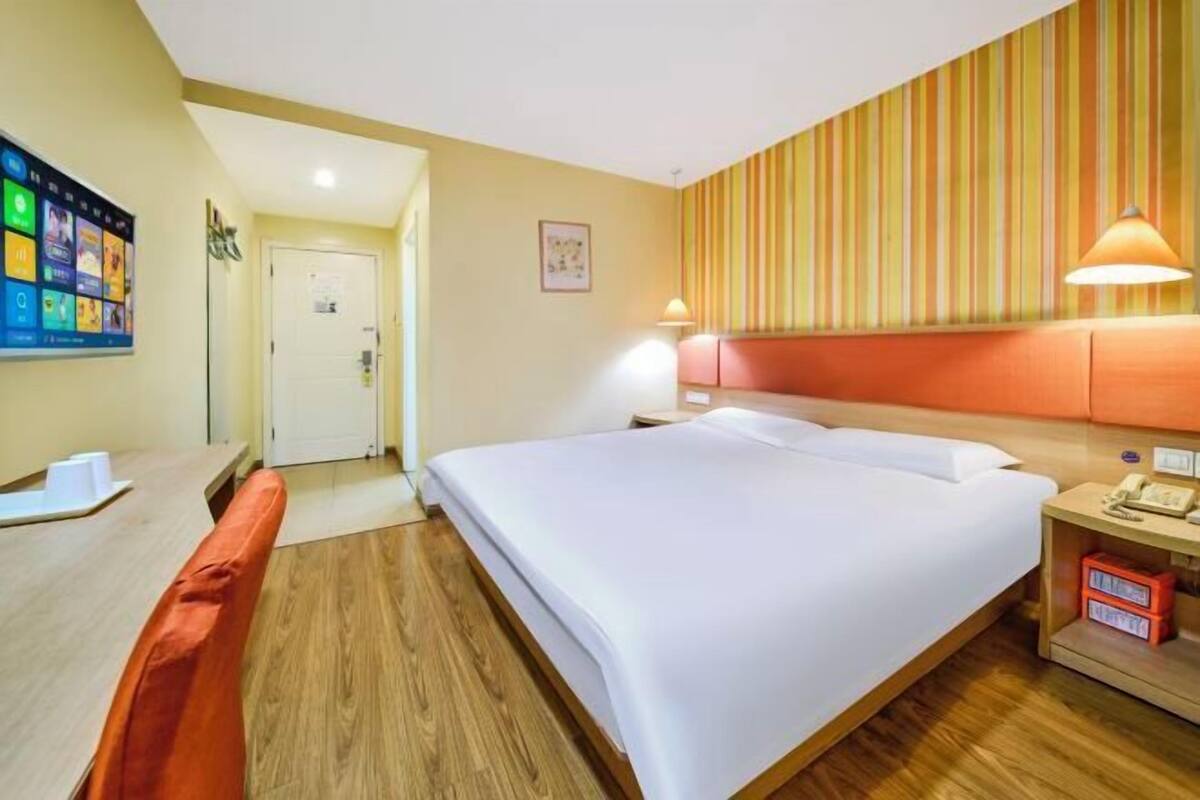 Habitación de hotel con una cama grande, un escritorio con computadora, una silla, una mesita de noche con lámpara y un televisor montado en la pared.