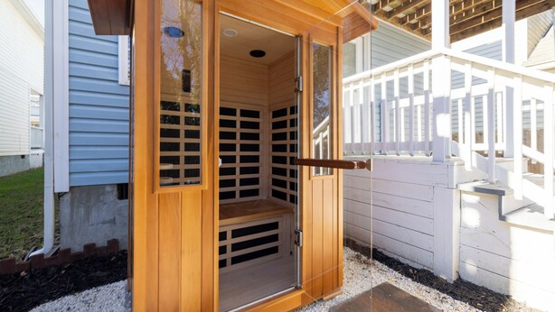 Una sauna de madera con un banco en el interior, una puerta de madera y una valla blanca al fondo.