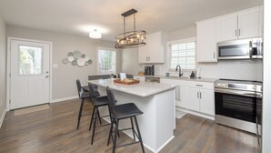 Una cocina moderna con armarios blancos, una isla central con taburetes de bar, electrodomésticos de acero inoxidable y una lámpara colgante.
