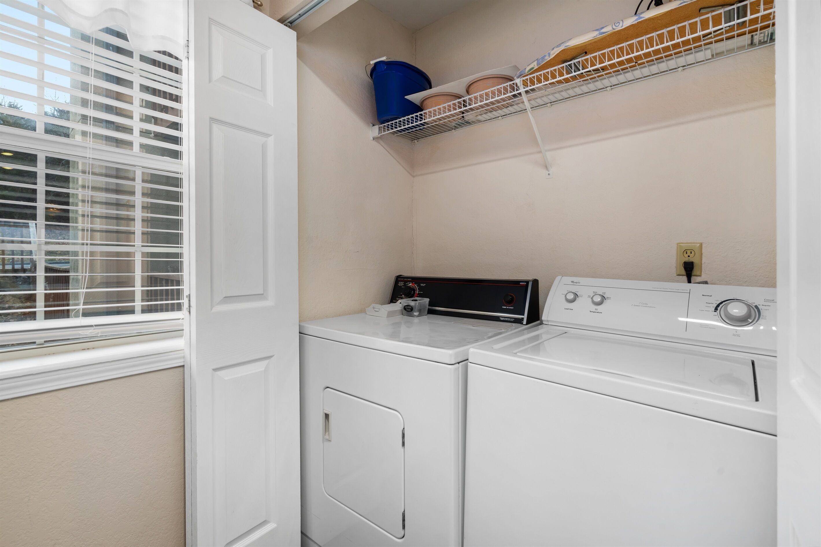 A laundry room with two white front-loading washers and dryers, a window with blinds, and a shelf above the dryers holding a blue bucket and potted plants.