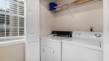 A laundry room with two white front-loading washers and dryers, a window with blinds, and a shelf above the dryers holding a blue bucket and potted plants.