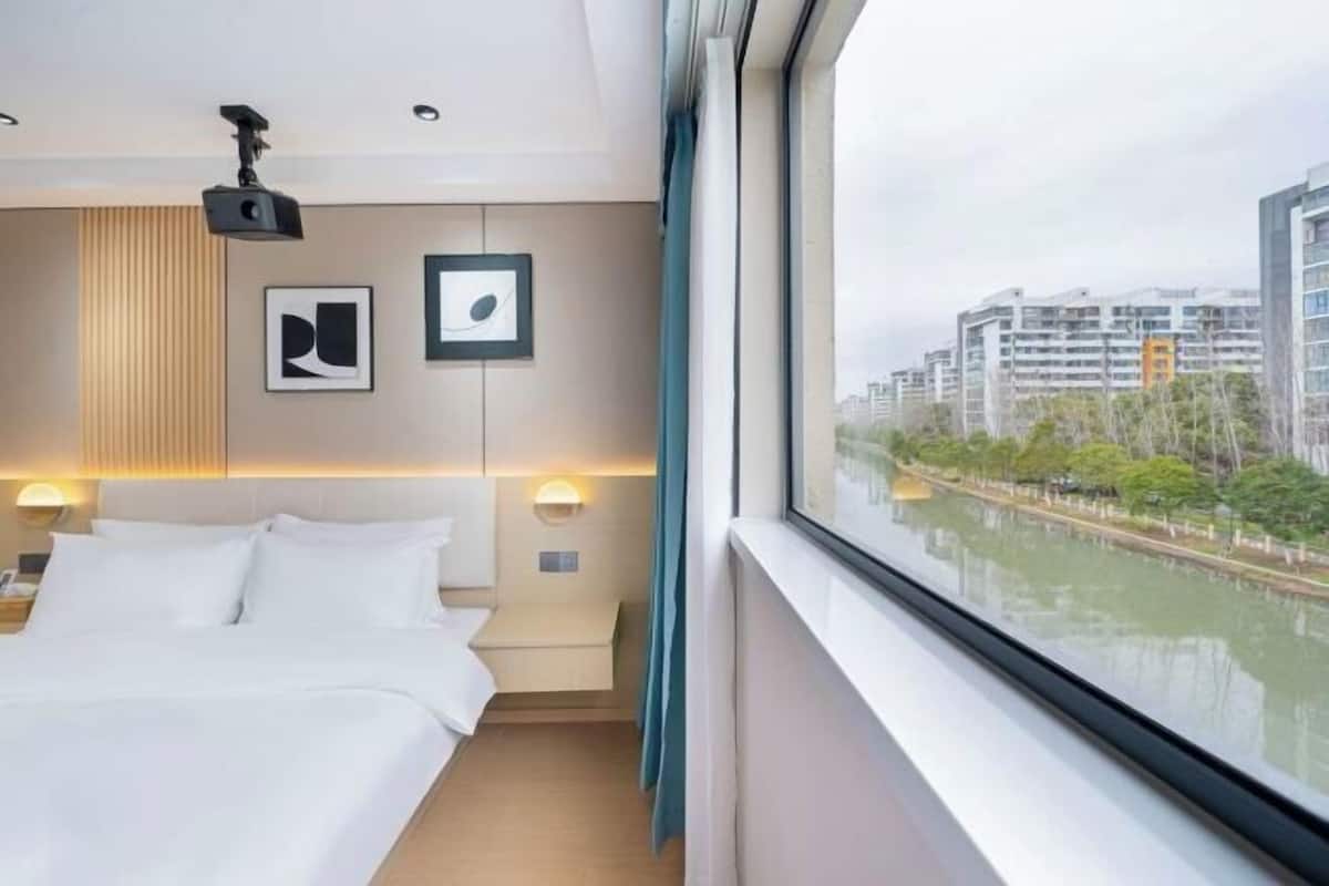 A hotel room with a bed, a projector, framed artwork, a nightstand, and a view of a river and buildings.