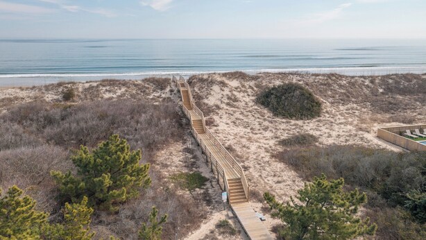 Ein hölzerner Steg führt zu einem Strandbereich mit Dünen und spärlicher Vegetation.