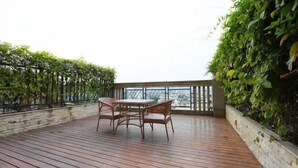 A wooden deck with a table and chairs, surrounded by lush greenery and a stone wall.