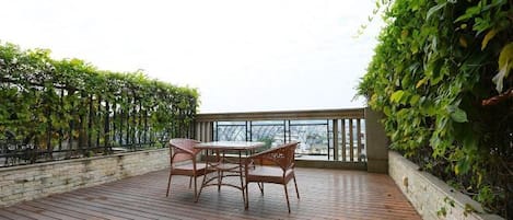 Una terraza de madera con una mesa y sillas, rodeada de follaje y una barandilla de vidrio.