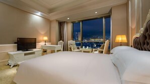A hotel room with a large bed, a desk, a chair, a television, and a view of the city at night.