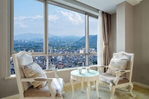 A room with two white chairs and a small table, large windows with a city view, and a curtain on the right. - Huidu Hotel (Longmen)