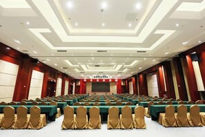 Uma sala de conferências com mesas e cadeiras dispostas em fileiras, um palco e uma tela grande.