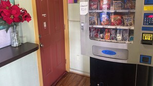 A vending machine with a 'PUSH' button and a coin slot, located next to a red door and a vase of red flowers.
