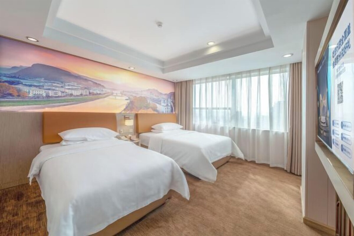 A hotel room with two beds, a large mural on the wall, and a view of a cityscape through the window.