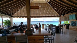 An outdoor dining area with wooden tables and chairs, a bar counter, and a view of the sea.