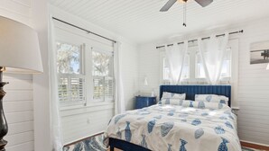 Un dormitorio con cama, ventilador de techo, ventana con cortinas blancas y suelo de madera.