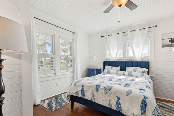 A bedroom with a bed, a ceiling fan, a window with white curtains, and a wooden floor.
