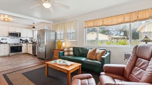 Una sala de estar amplia con un sofá verde, un sillón de cuero y una mesa de centro de madera. Se ve una cocina con armarios blancos y electrodomésticos de acero inoxidable al fondo.